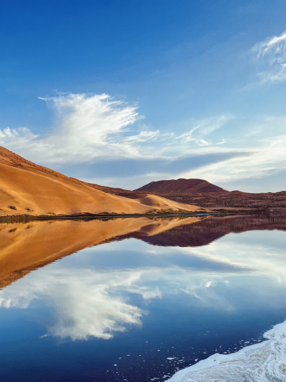 Blue lake mirroring the sky beside a dune slope and red-brown hills at Tianchi