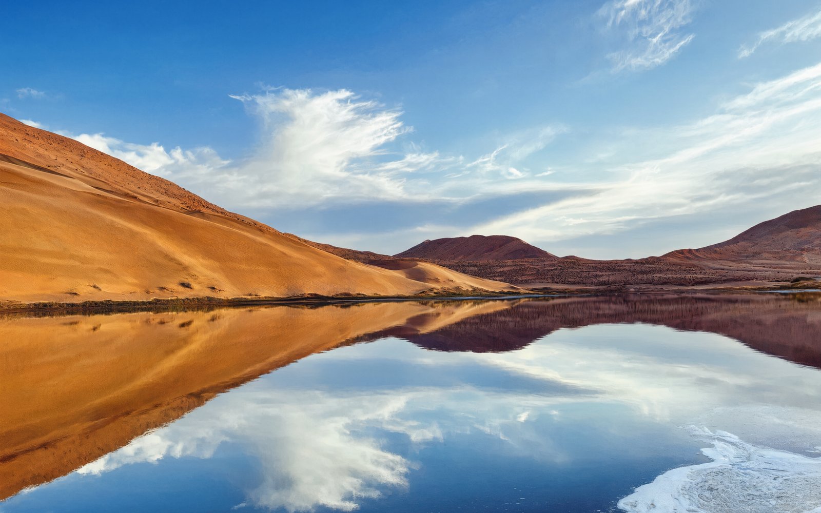 Blue lake mirroring the sky beside a dune slope and red-brown hills at Tianchi