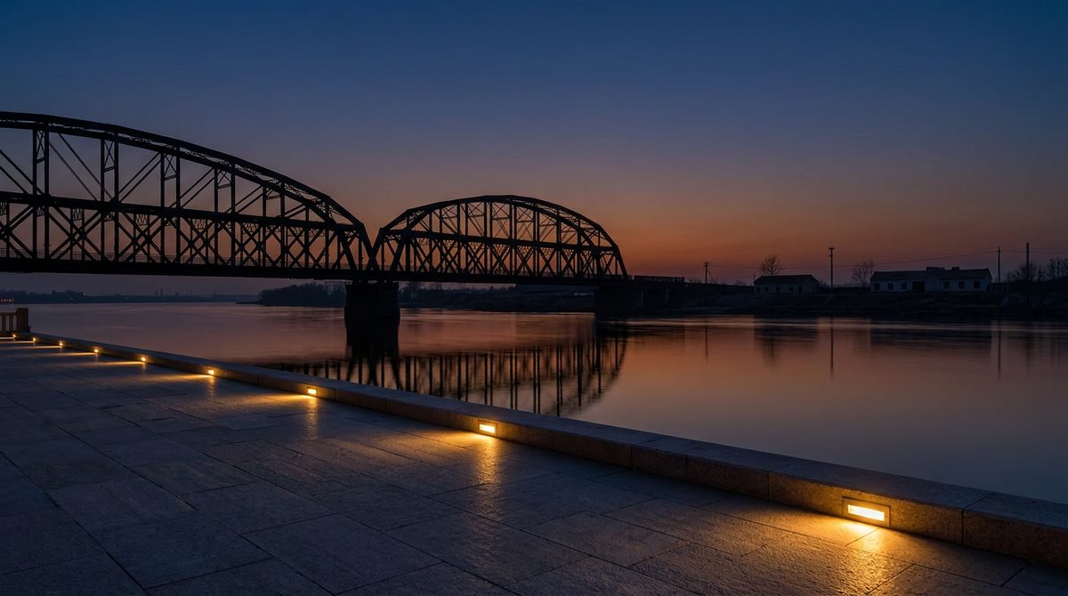 Dandong Yalu River promenade at dusk broken bridge silhouette over calm water with North Korean riverbank beyond warm lights
