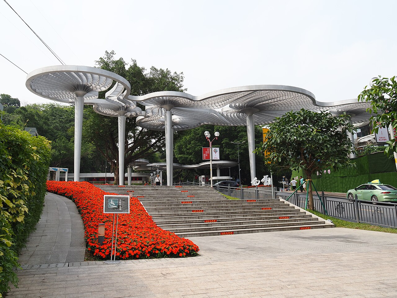 A set of grey mushroom-shaped metal canopies on tall stems over a paved plaza, with red flowers banked beside a flight of steps