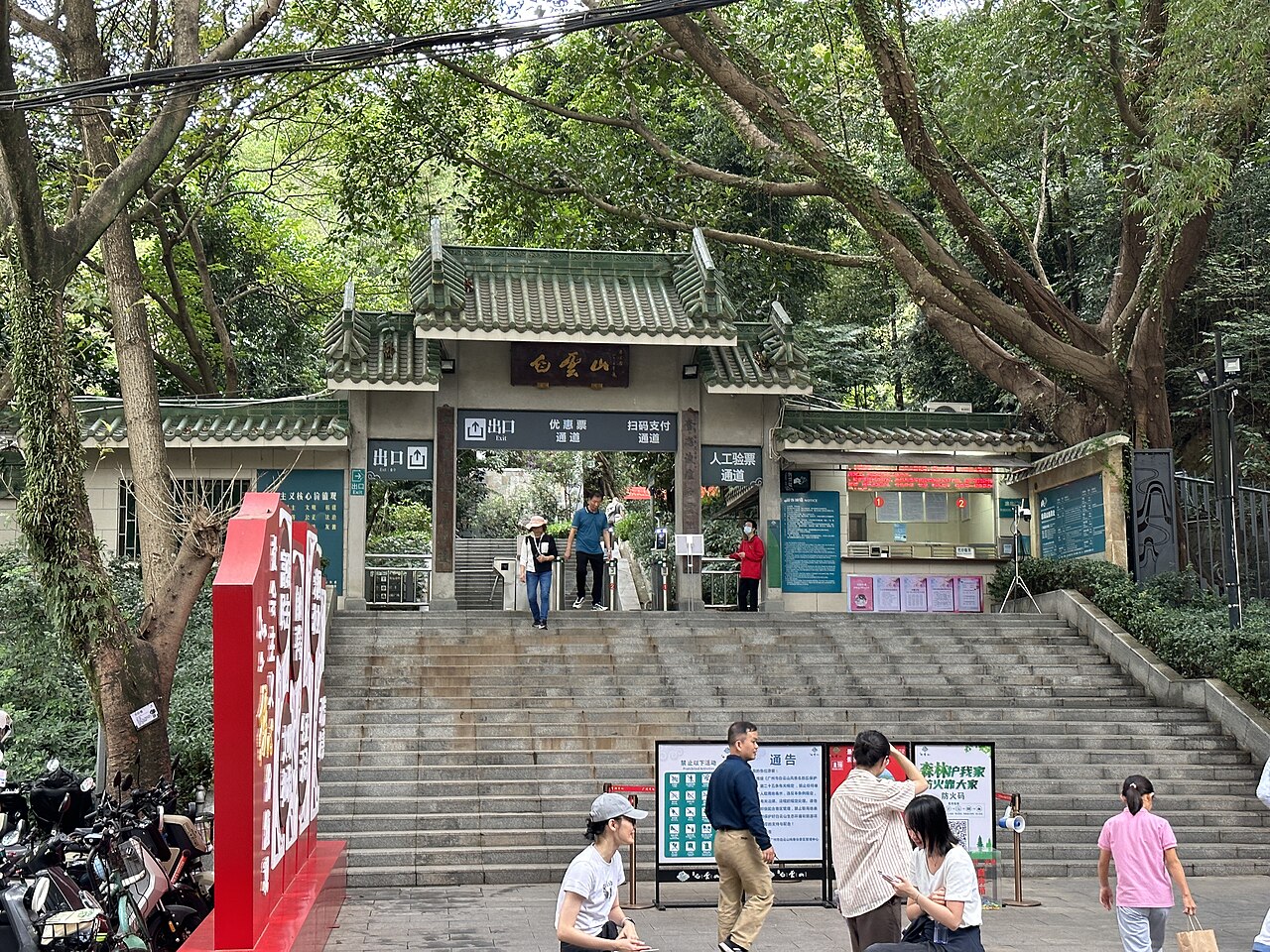 A green-tiled gate building under large old trees at the top of a wide flight of stone steps, with a few visitors on the steps