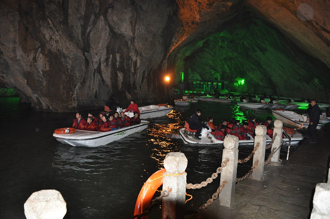 Several boats carrying visitors in red jackets moving along a dark underground river beside white floats and rails, under a rock ceiling with green light on the far wall
