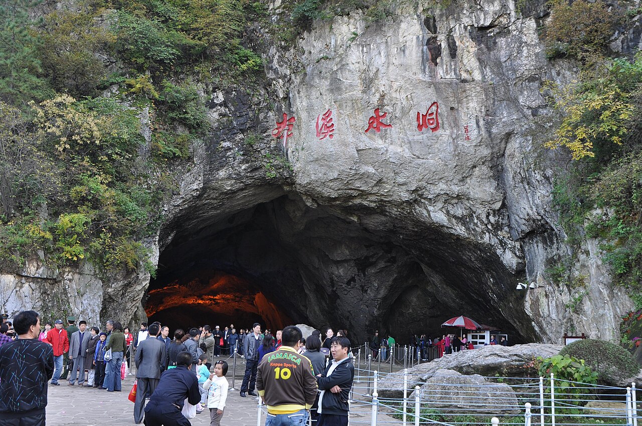 A wide cave mouth in pale limestone with large red characters carved above it, with trees on the rock and a crowd standing on a paved forecourt in front