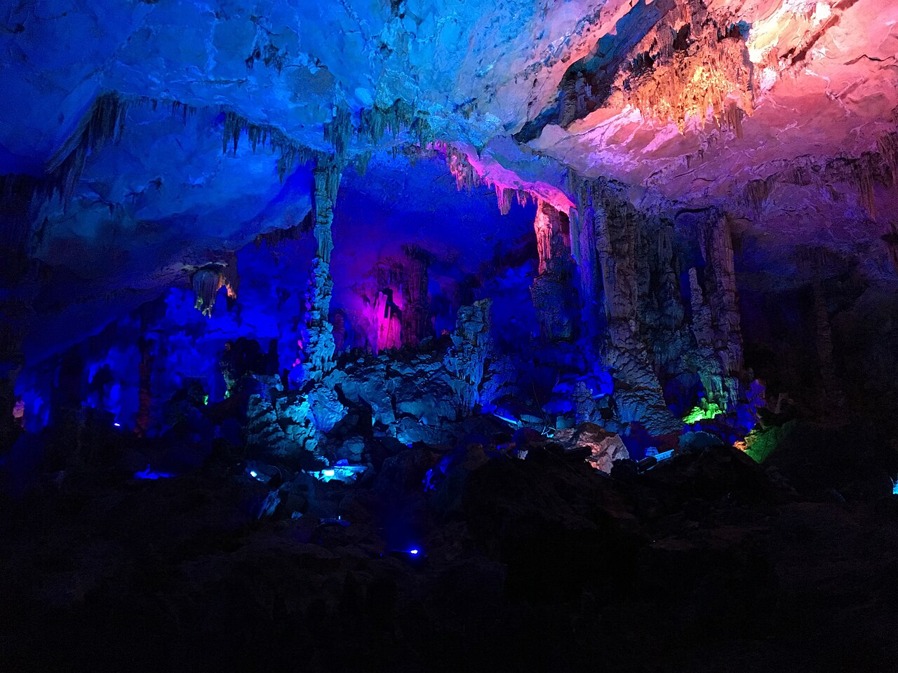 Cave formations lit in blue, pink and green, rising from dark water under a rock roof