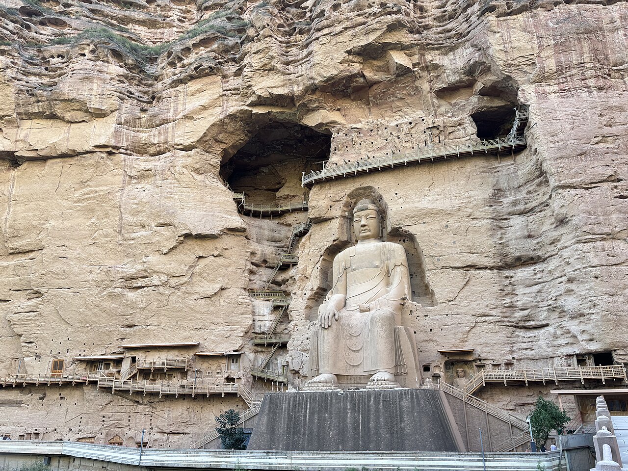 The seated Buddha in its cavern with a ring of cave openings above and wooden walkways winding up the cliff