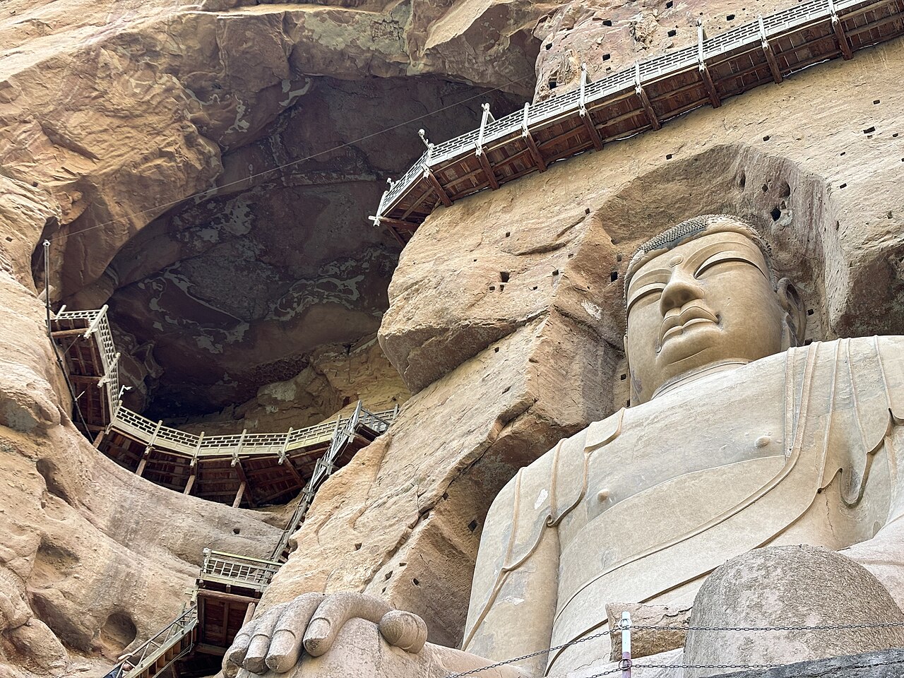 The face and shoulder of the large seated Buddha with a long wooden walkway curving along the rock above, in a dark cavern