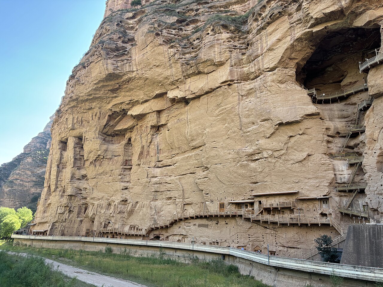 A broad pale sandstone cliff face with dark cave openings along it and a path and green grass at its foot