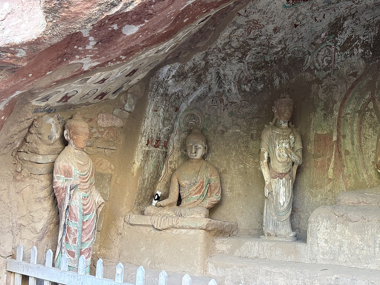 A cave niche with a seated Buddha between two standing figures, in warm stone with traces of red and green pigment, behind a white railing
