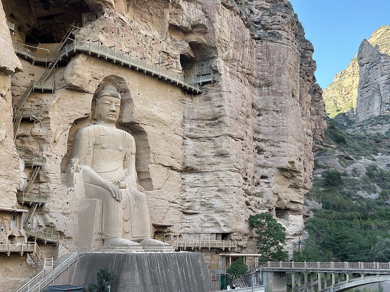A huge seated Buddha carved in a sandstone cavern in a cliff, with a wooden walkway and railings at its base and a blue patch of sky to the right