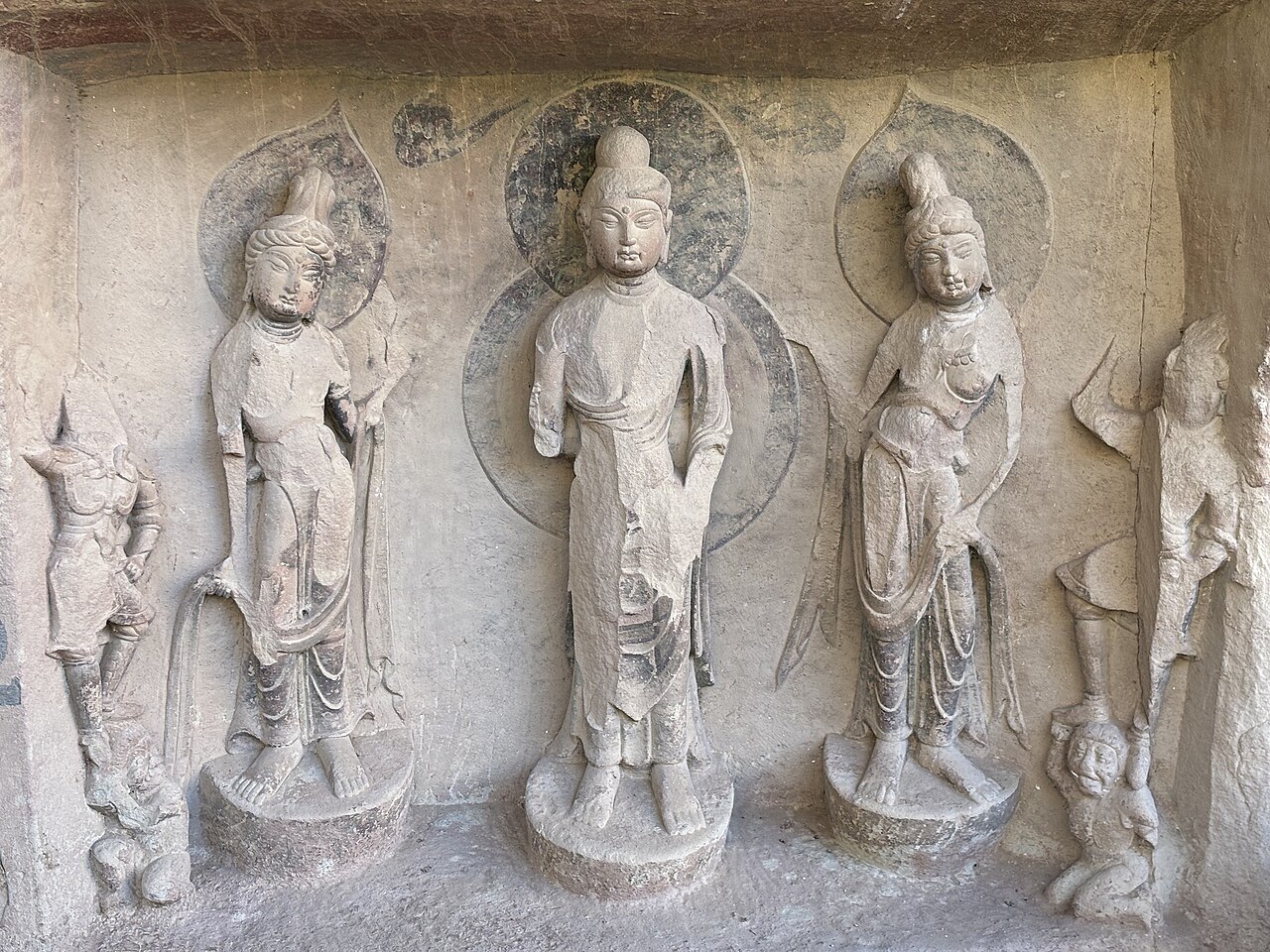 Three tall standing figures carved in relief in a stone niche, the central one with a halo, with small figures at the sides