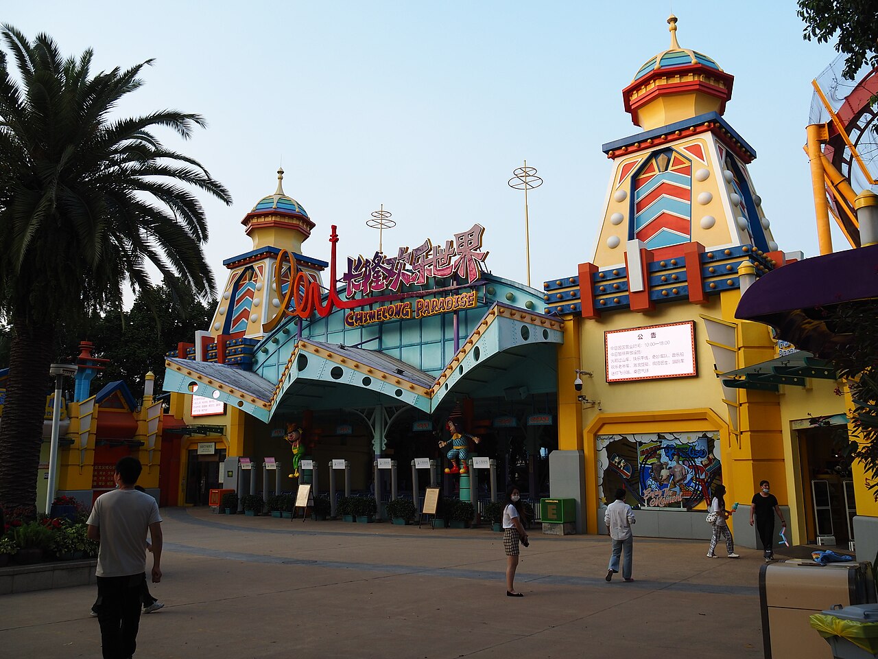 The colourful main gate of Chimelong Paradise with towers topped by golden domes, bright painted facades and palm trees, with a few visitors walking on the plaza in front