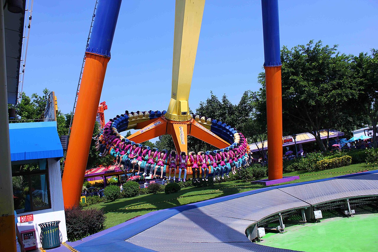 A giant pendulum ride with tall orange and blue supports, with a ring of seats at its base and green trees around