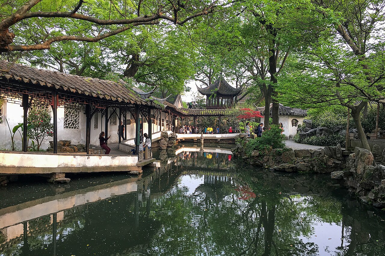 A covered walkway with a tiled roof and red columns running along the pond edge in spring green, with a pavilion and trees beyond