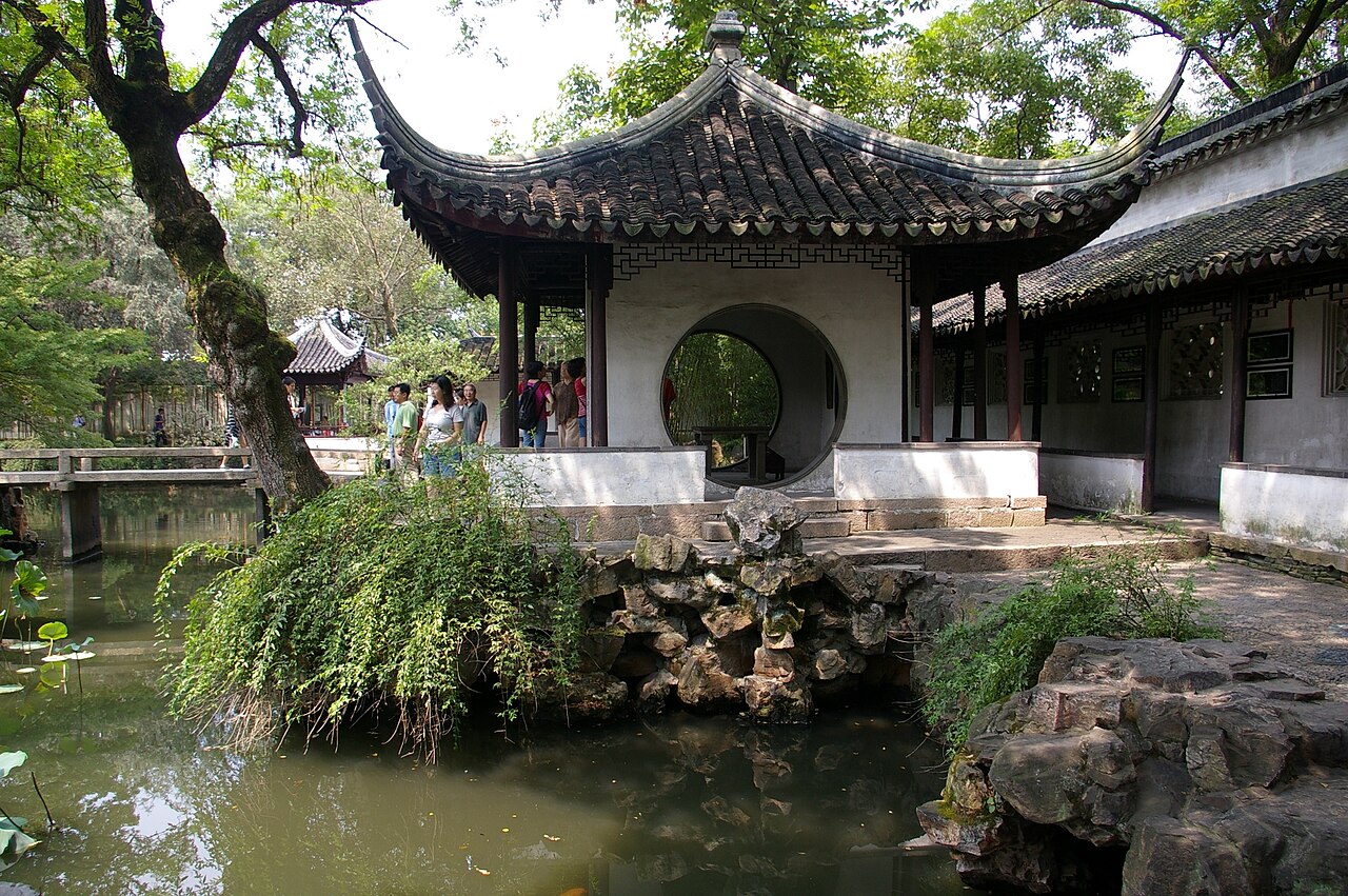 A white-walled pavilion with a round moon-gate opening and a curved tiled roof, set beside a pond with rocks and ferns in the foreground