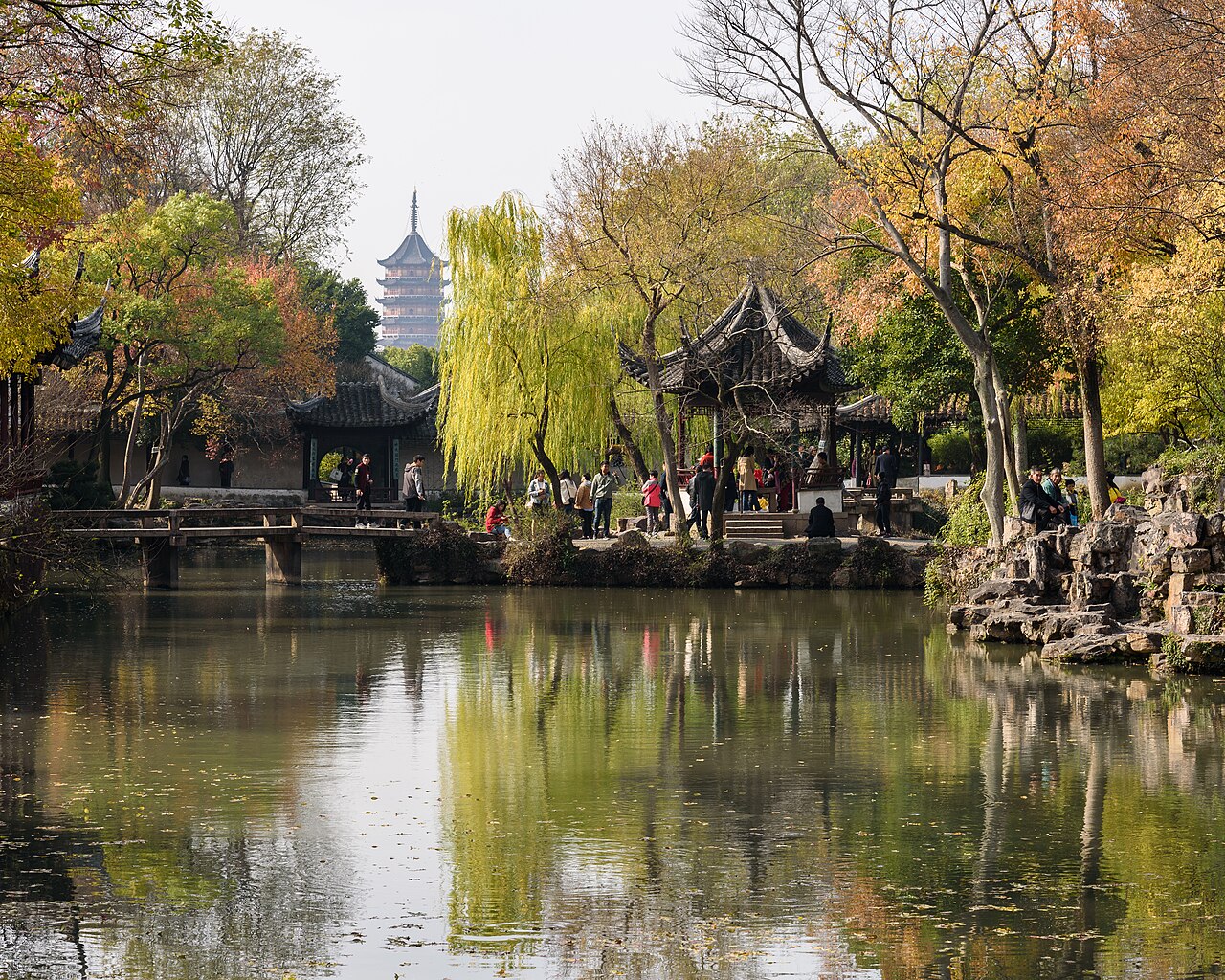 Across a pond lined with autumn trees and a rock bank, a small pavilion and a distant pagoda rise above the trees