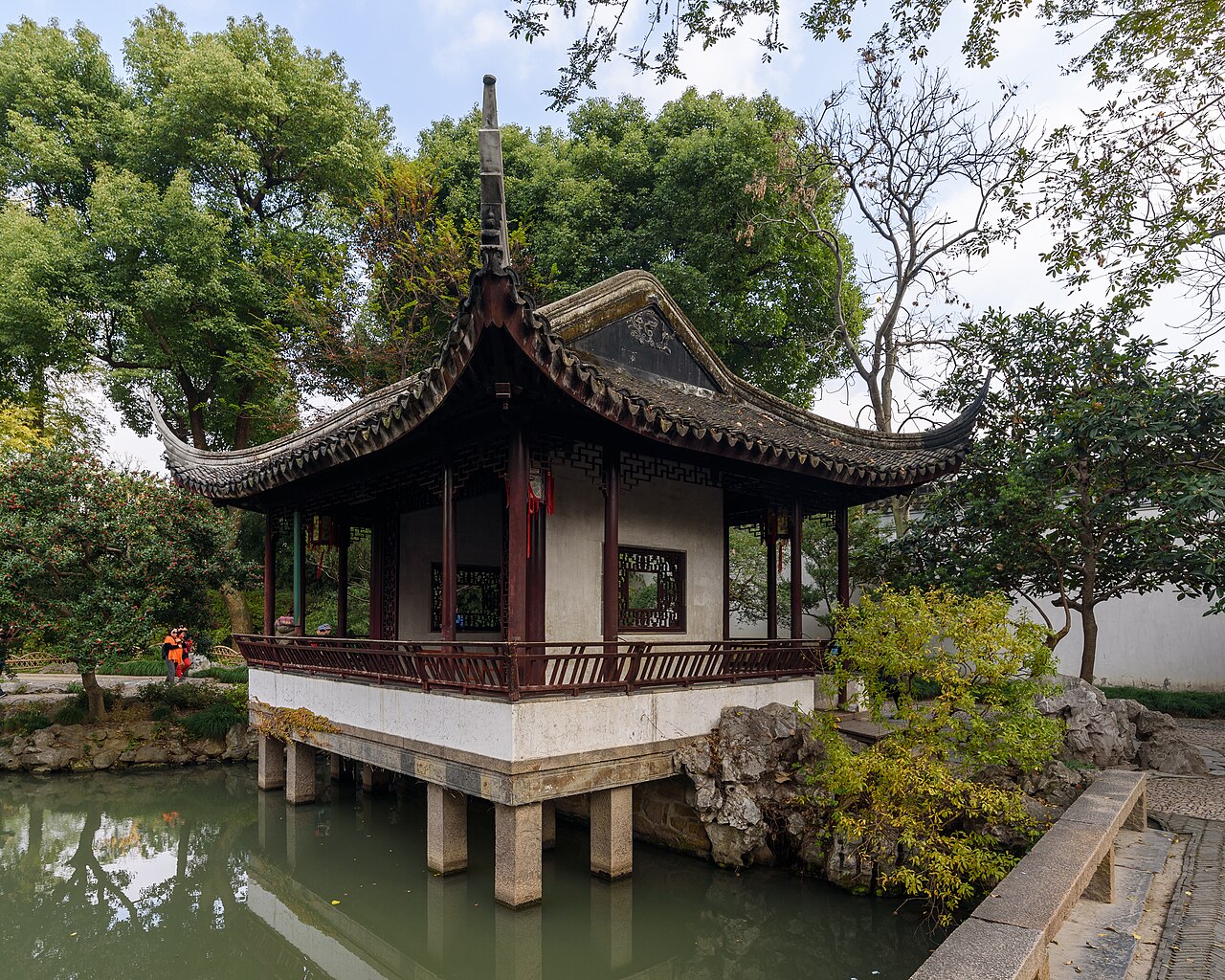A small dark-roofed pavilion with red columns standing on stone piers at the edge of a pond, with a white wall and trees behind