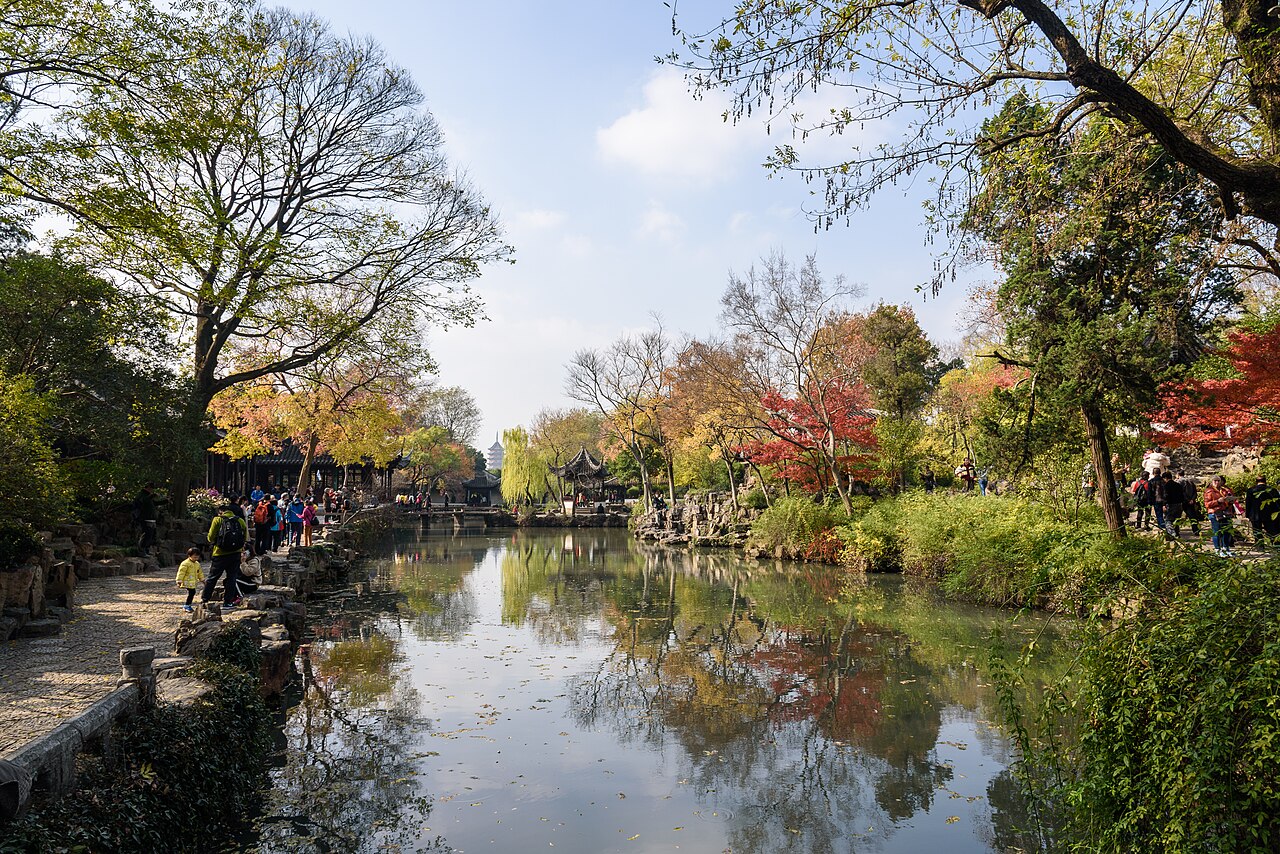 A broad pond in a Suzhou garden with autumn-coloured trees along the far bank, a stone path at the left and pavilion roofs among the branches under a pale blue sky