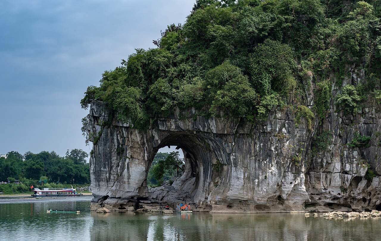 The same limestone hill from a nearer angle, with the round arch at the waterline, a tree-covered crown and a river boat in the distance