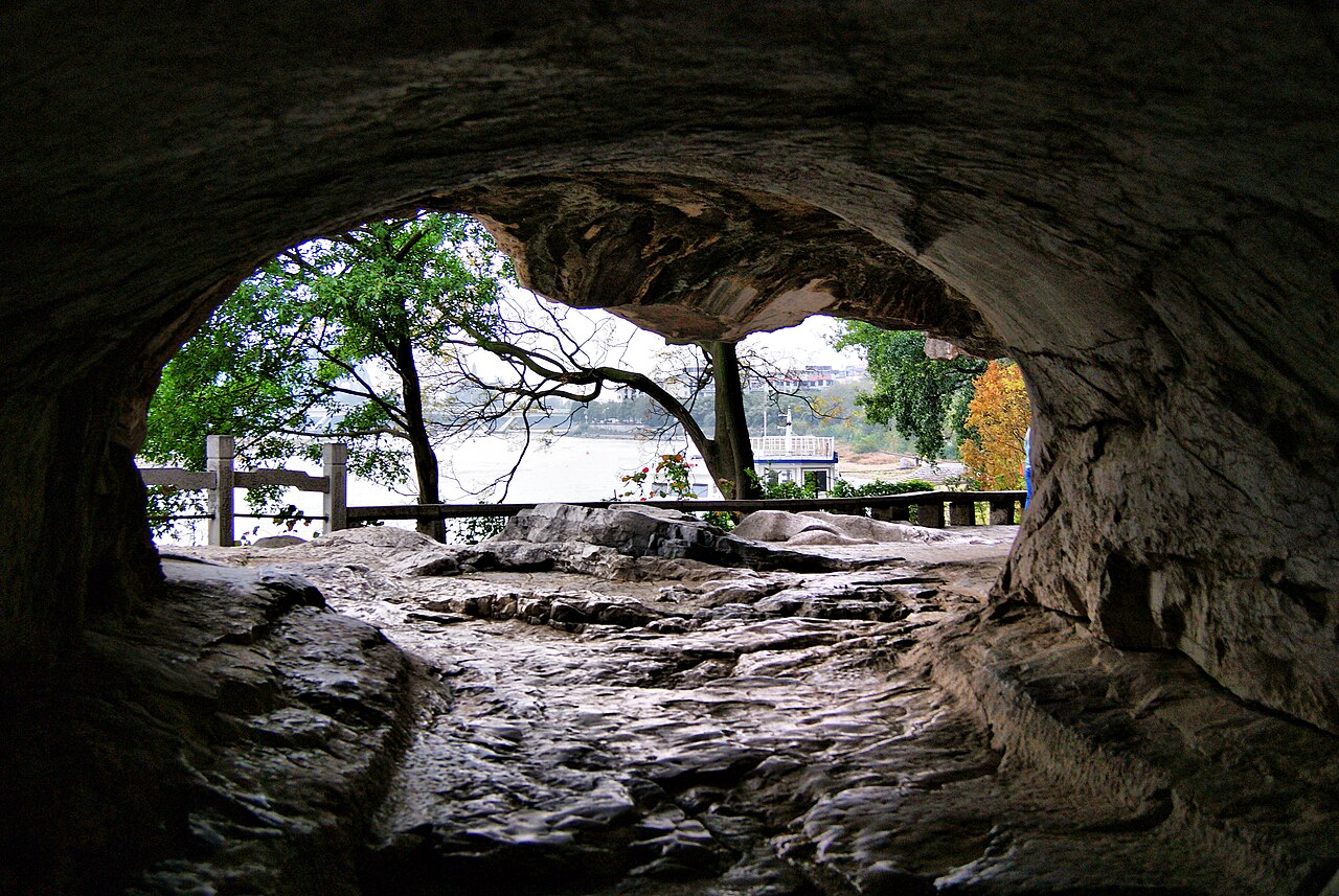 Looking out from inside a limestone cave through a rough opening to trees, a railing and a river beyond