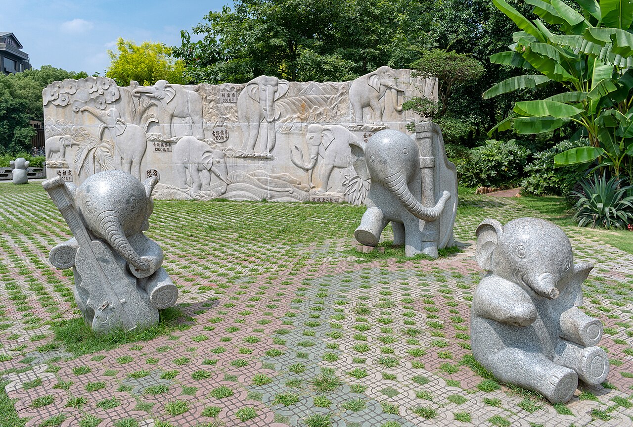 A paved court with stone sculptures of baby elephants in front of a long carved stone relief wall, with a large leaf plant at the right