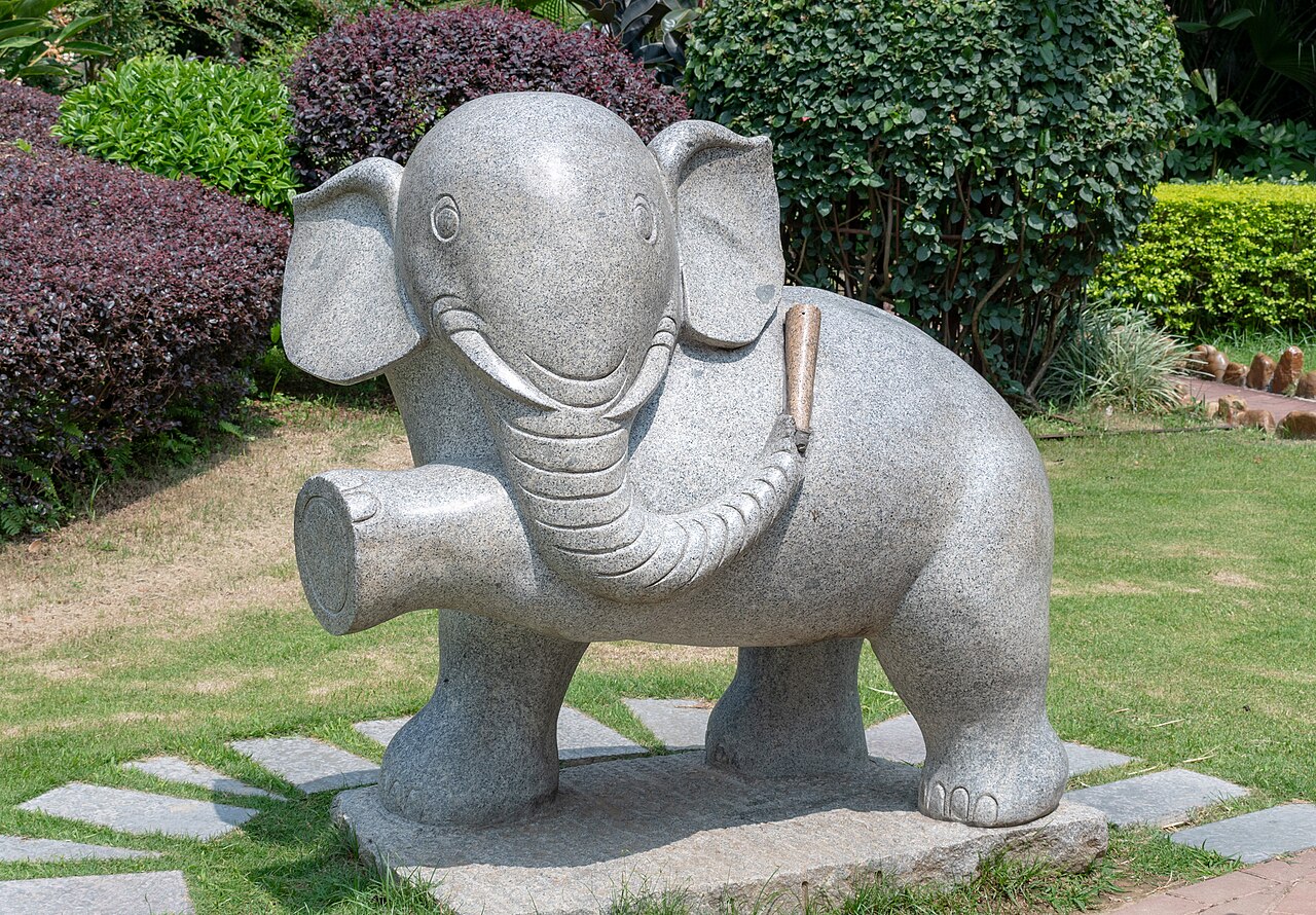 A grey granite elephant statue with a raised front foot standing on a lawn bordered by low clipped shrubs and stone path slabs