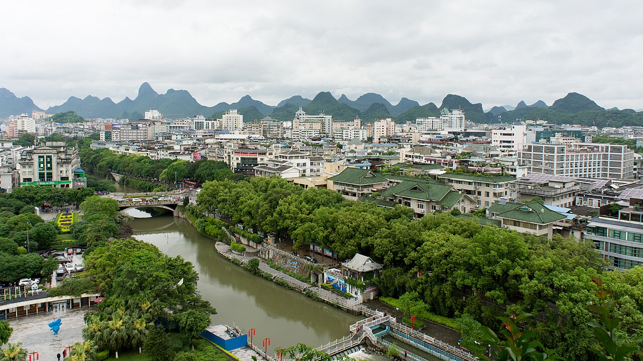 A view over the rooftops of Guilin with a green canal and trees in the foreground and a line of sharp limestone peaks on the horizon under cloud