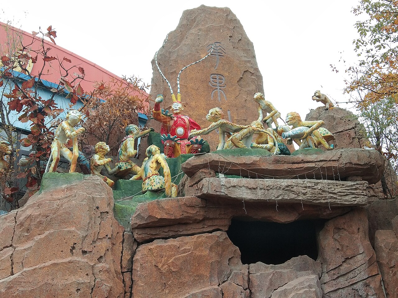 A group of painted statues of monkeys clambering over a large rock with red characters and a dark cave opening below, with a temple roof at the left