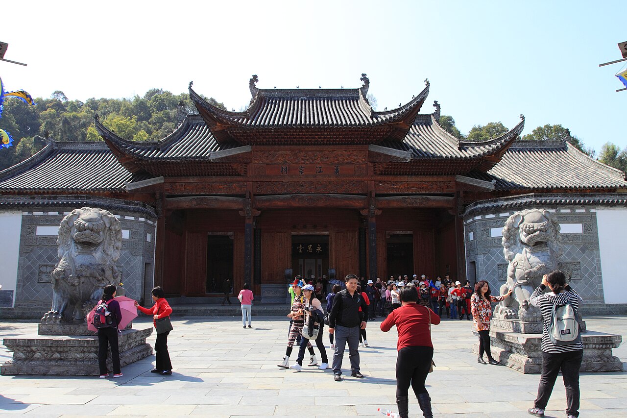 The front of a village ancestral hall with a layered curved roof ridge, a pair of stone lions on either side of the step and a group of visitors in the court in front, with wooded hills behind