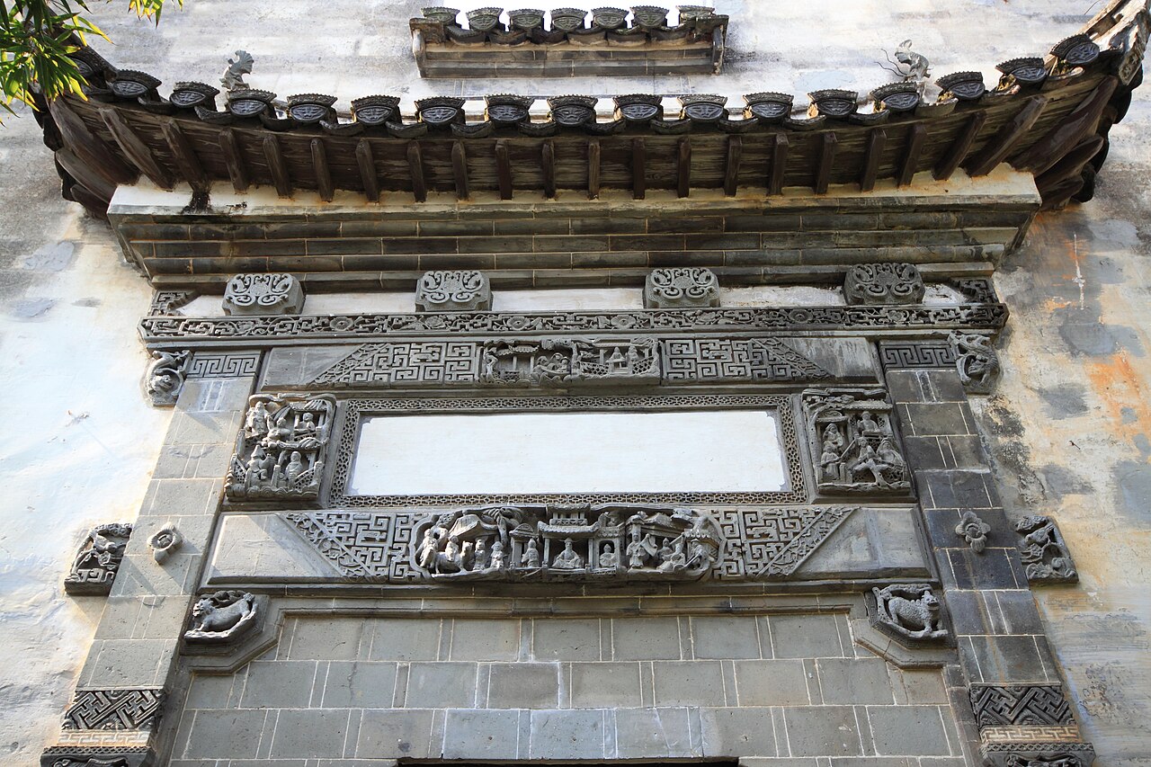 A carved brick gateway with layers of relief figures and floral panels under a tiled eave, set into a pale wall