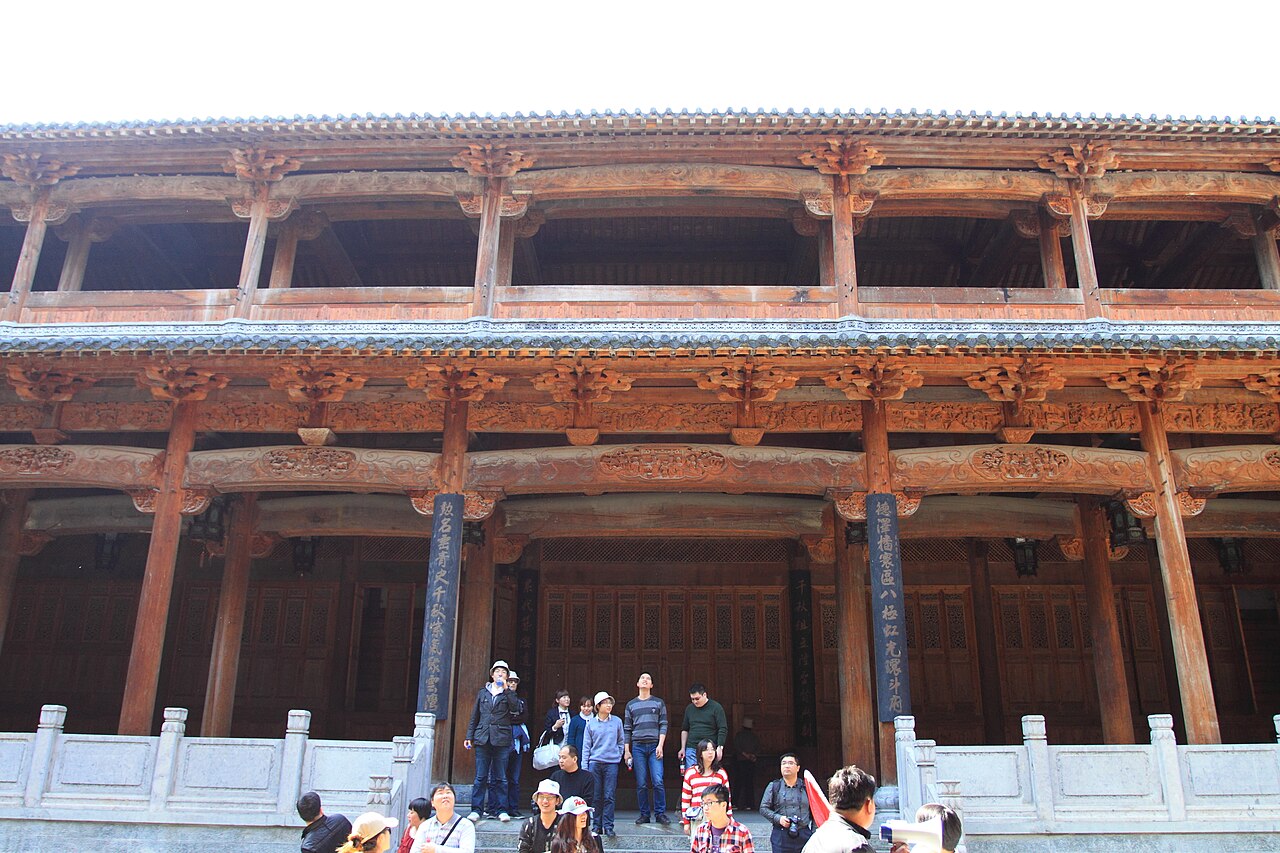 The open timber front of a hall with a carved wooden gallery along its upper storey, grey stone balustrades on the platform and a small group standing at the foot of the steps
