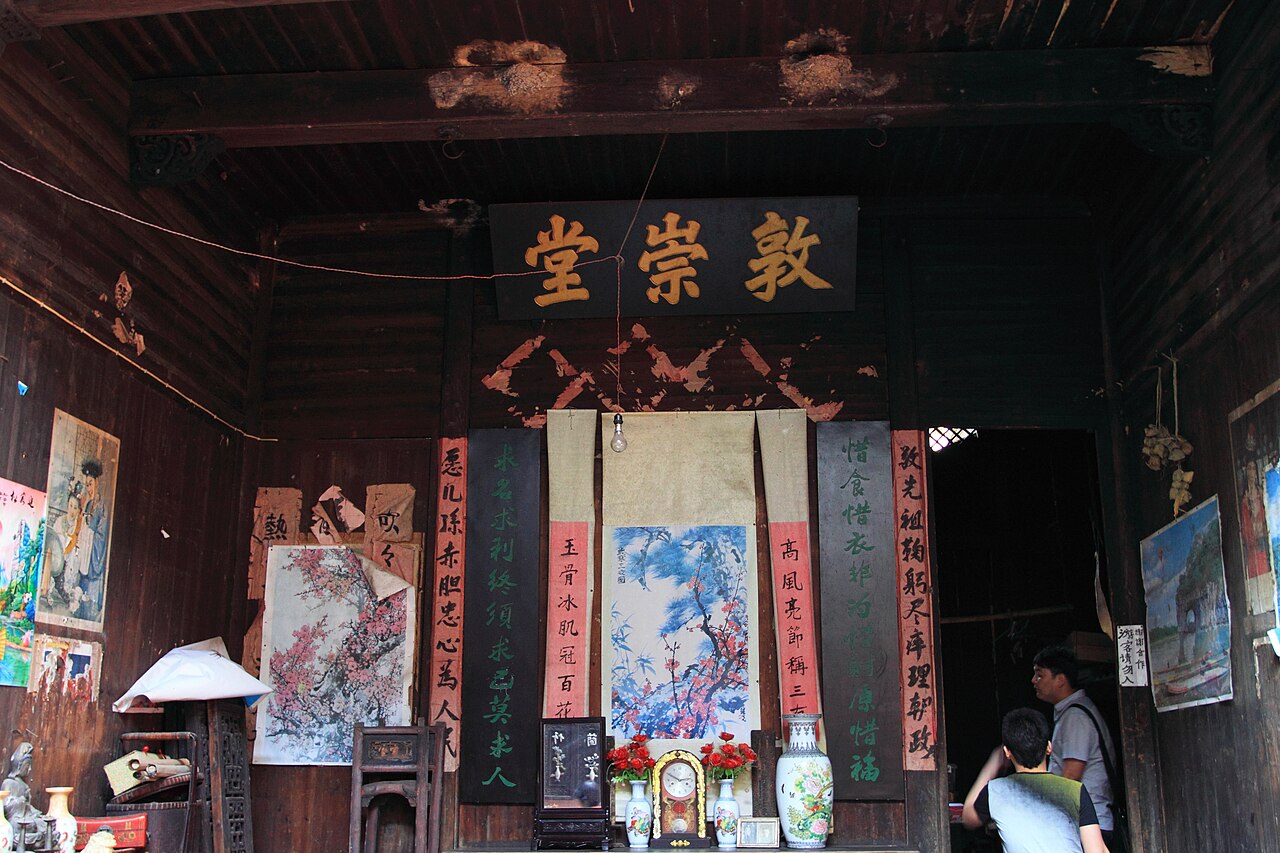 Inside a timber hall with a dark ceiling, a name board hanging above the doorway, red couplets on the pillars and painted panels leaning against the wall