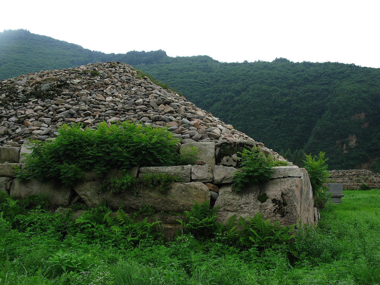A mound of rough grey stone with a stepped base of cut blocks, partly covered by green plants, with forested mountains rising behind