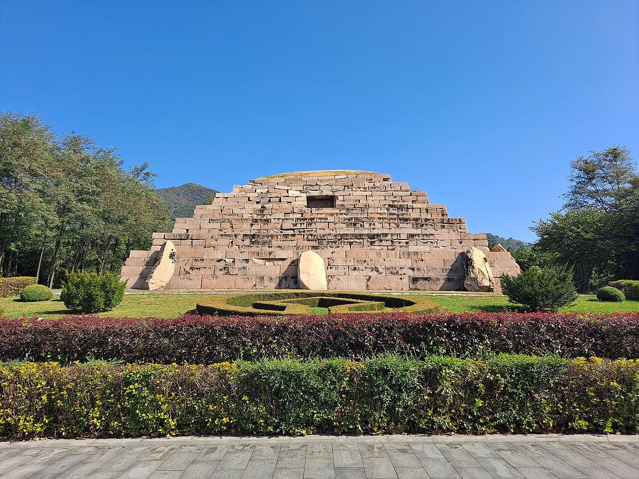 A flat-topped stepped stone tomb on a lawn beyond a long bed of flowering shrubs, with a paved path in the foreground and low mountains behind