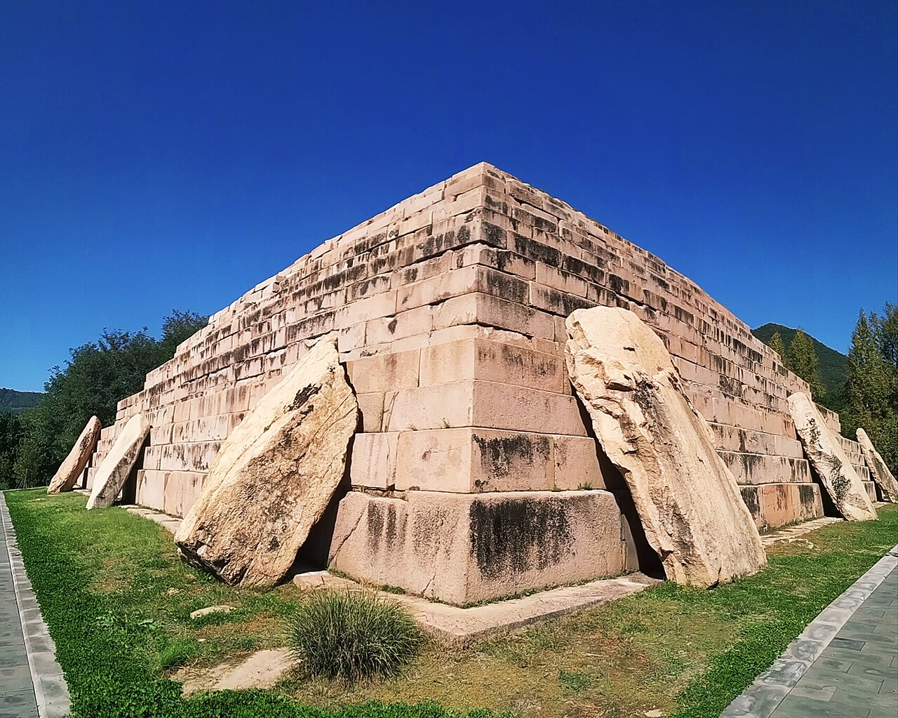 A stepped stone tomb seen from a corner, its tiers of cut blocks backed by large leaning slabs, against a deep blue sky