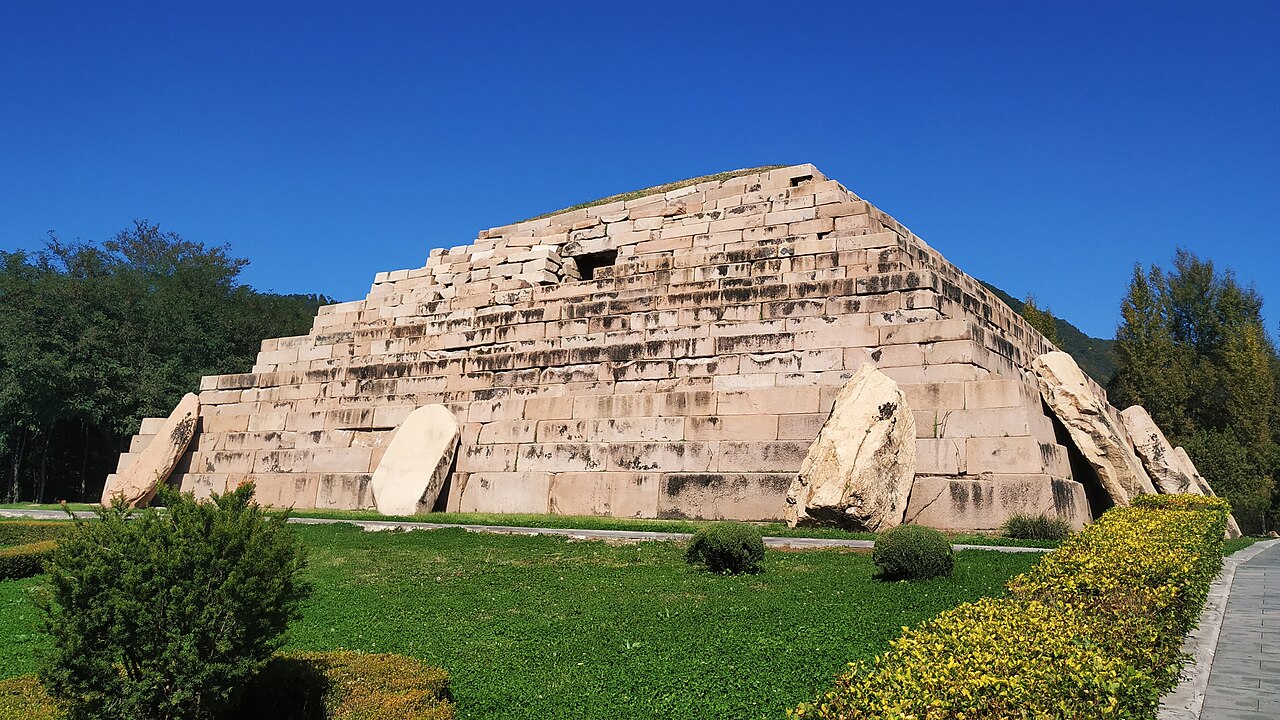 A stepped stone pyramid tomb of pale cut granite with large slabs leaning at its base, on a green lawn with clipped hedges and trees under a deep blue sky