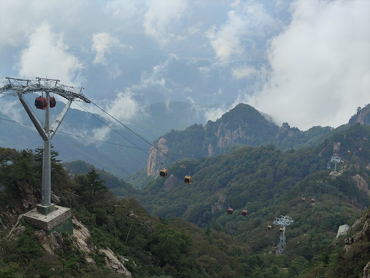 A steel pylon and cable carrying red cabins across a forested slope to a high granite ridge in the distance under broken cloud