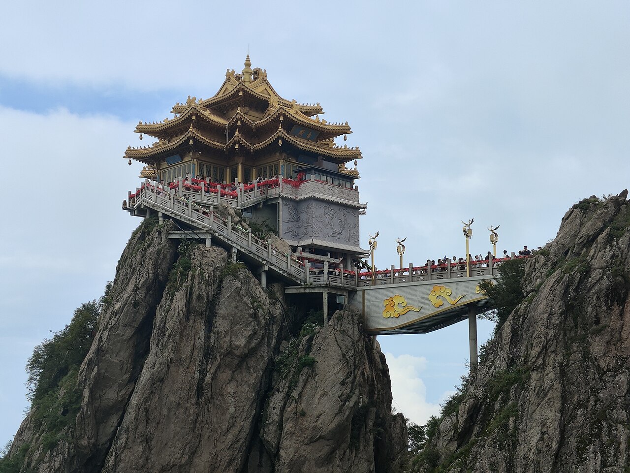 A timber pavilion with a double roof of gold-glazed tiles standing on a rocky spur, with a covered bridge leading to it and a few visitors on the railing beside it under a bright sky