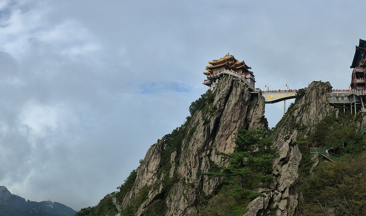 Two rock spurs with a small pavilion on the left one and a walkway bridge to a larger building on the right, with pines on the rock and thick cloud behind