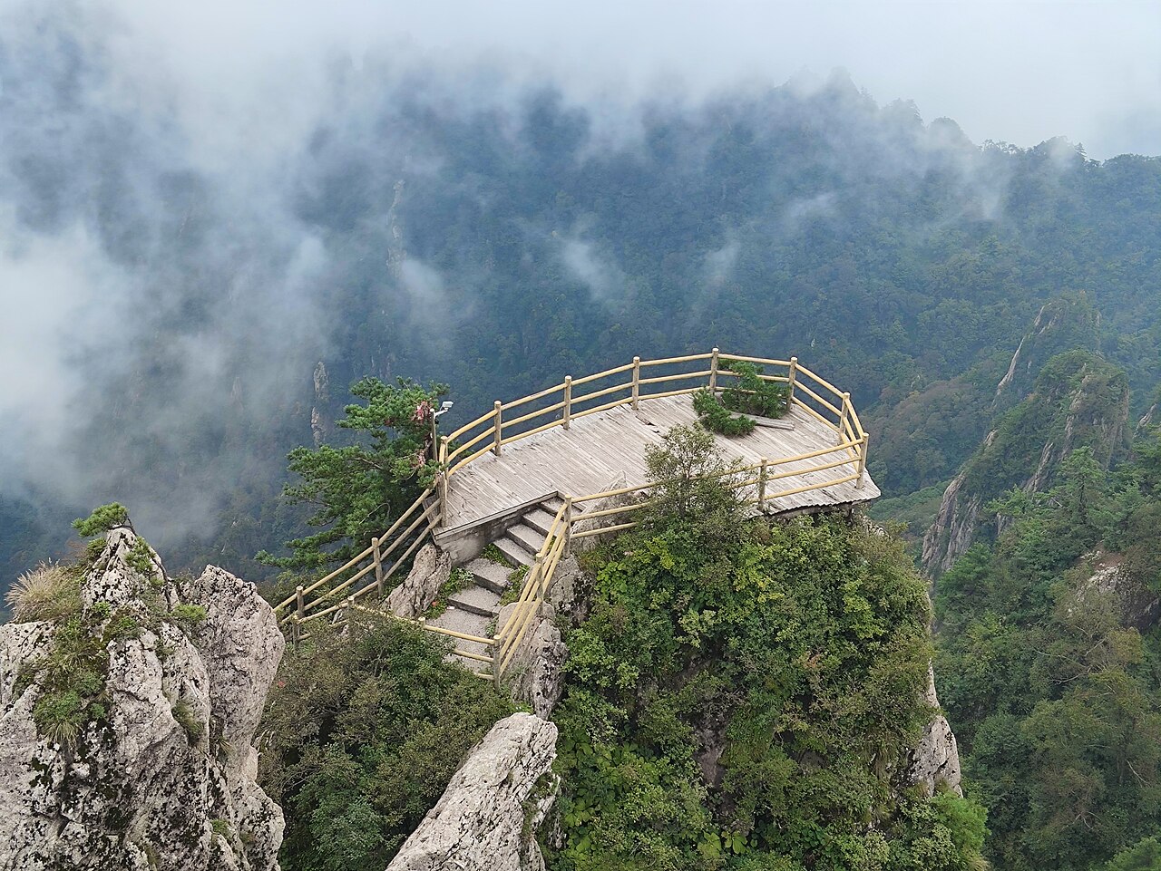 A curved timber viewing platform with a railing on a granite outcrop above a misty valley, with green slopes and low cloud behind