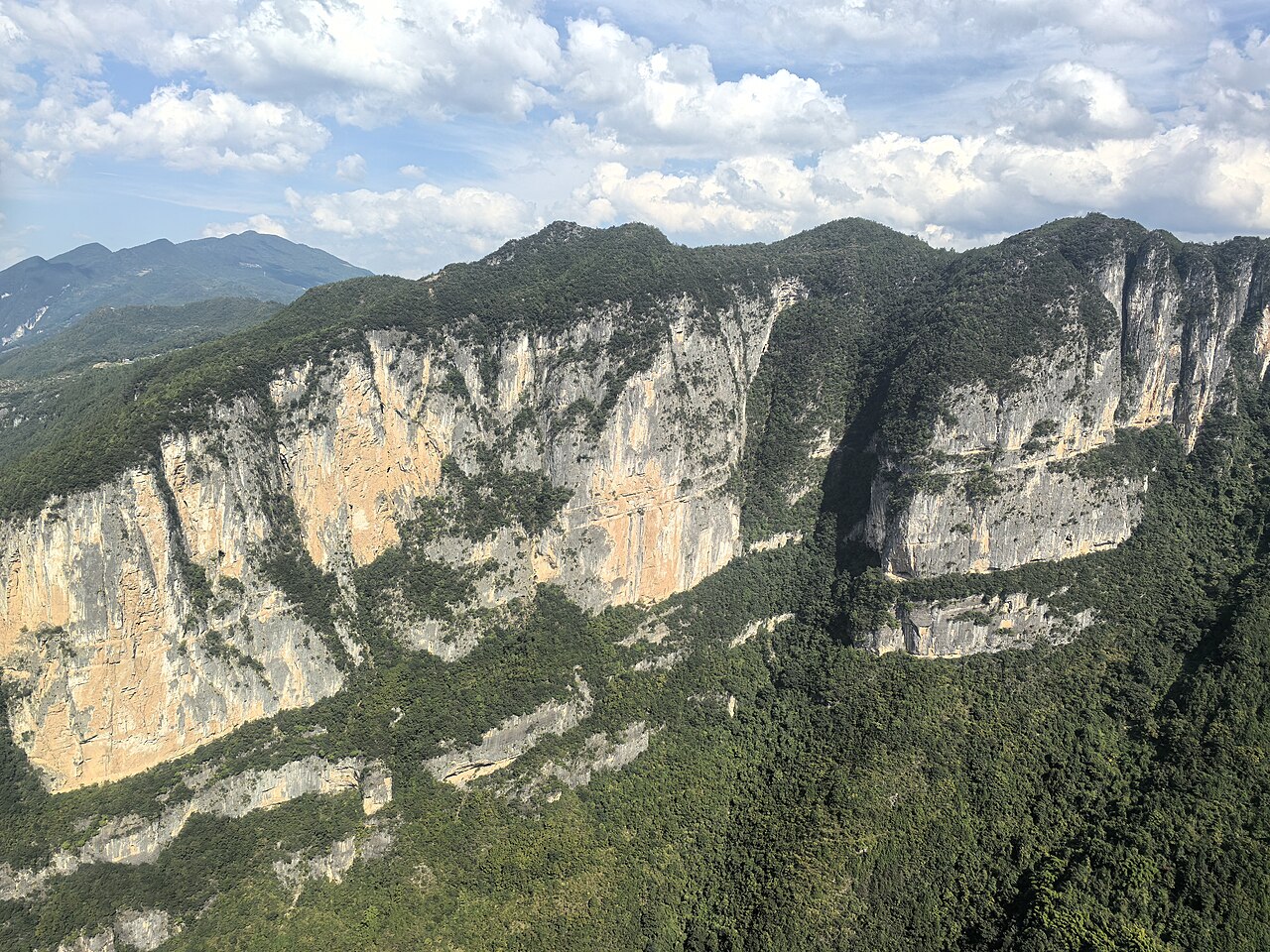 A long pale yellow limestone cliff band above a green forested slope, with blue mountains and cloud behind