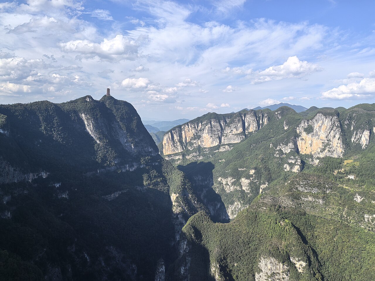 A deep gorge with a shaded wall on the left and sunlit limestone cliffs at the right, with a small watchtower on the ridge top at the left
