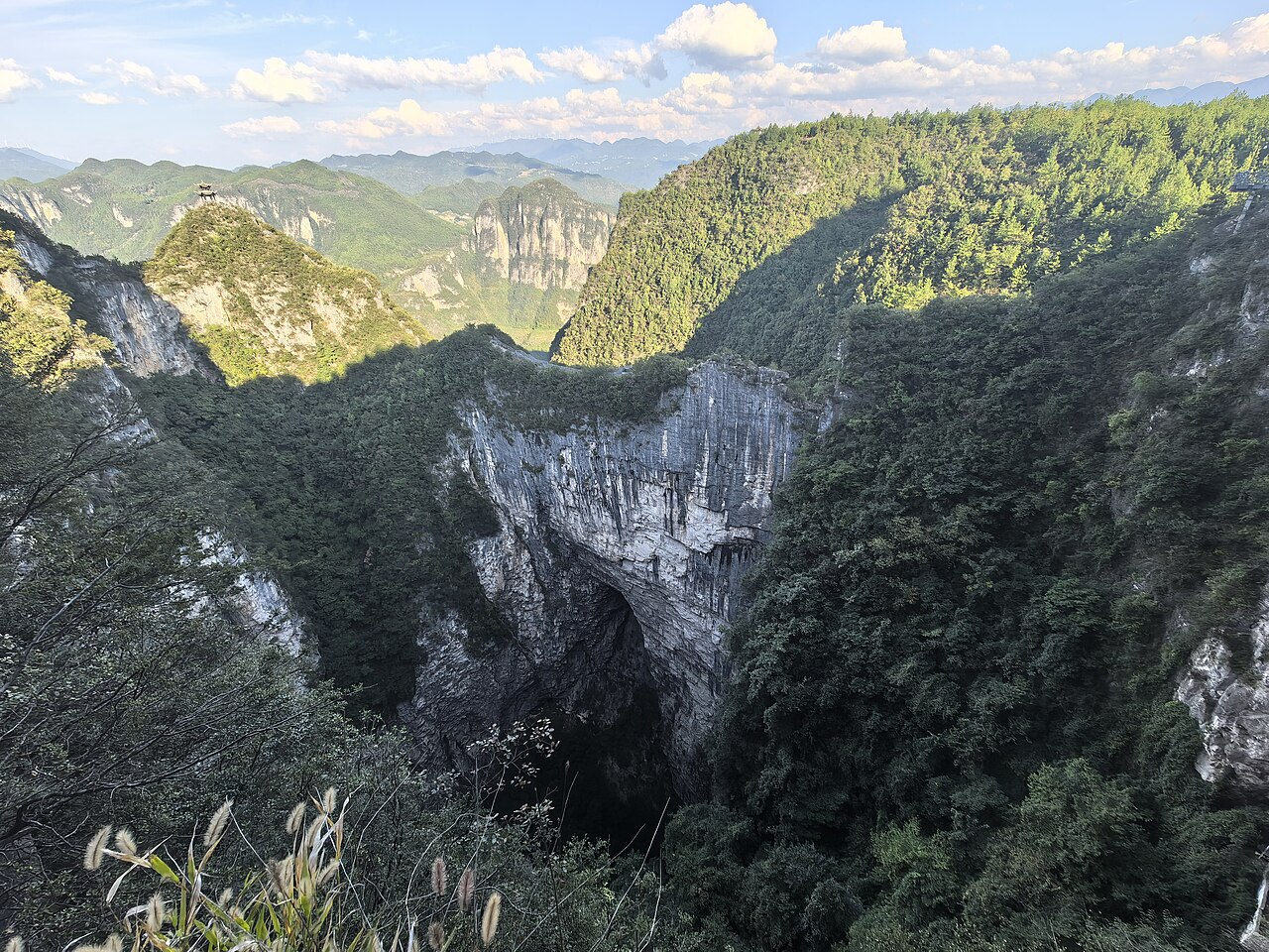 A deep rocky basin cut into a ridge with a pale limestone wall on the far side, trees on the near slope and layered mountains beyond