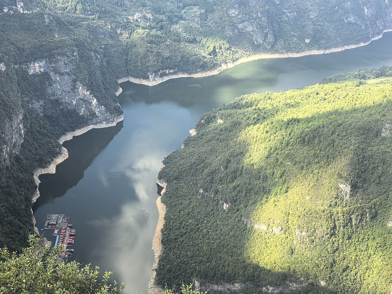 A river bend between forested ridges with a pale shoreline and bright green slope in the sun, seen from a high viewpoint
