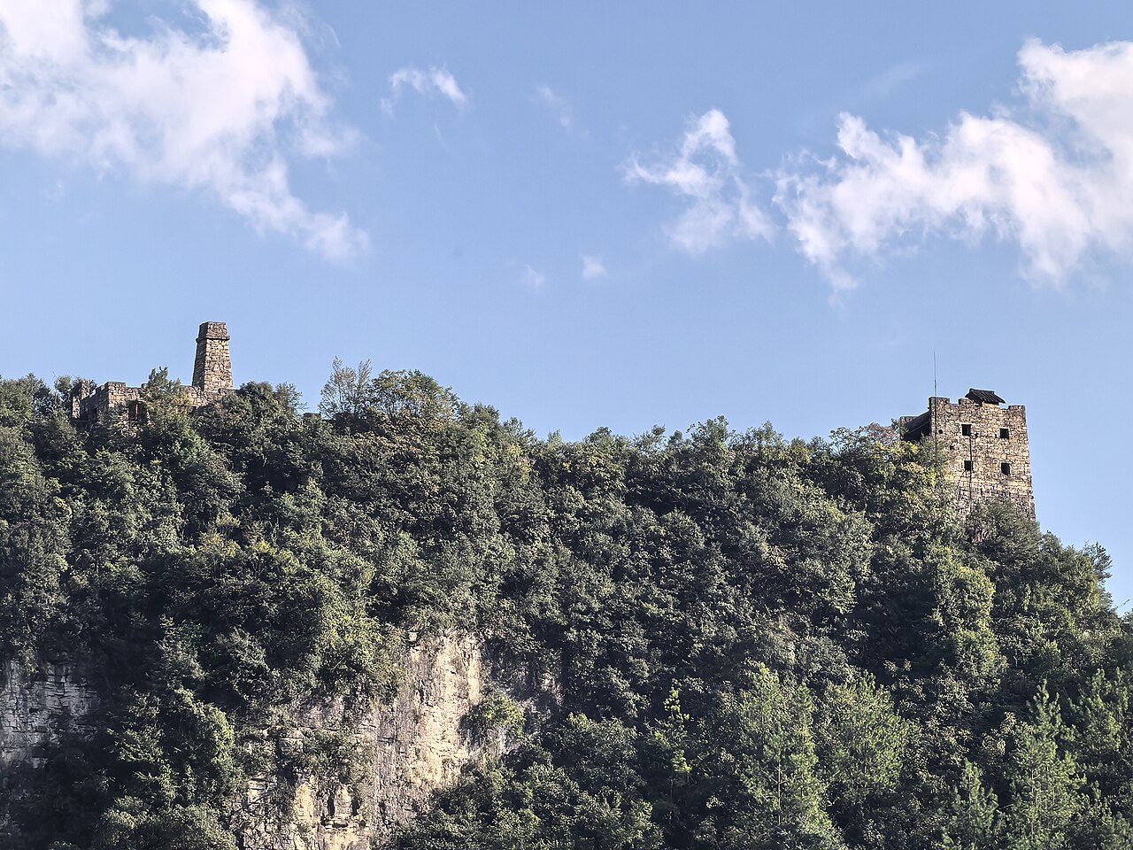 Two small stone watchtowers on the edge of a dark forested ridge against a blue sky with scattered clouds