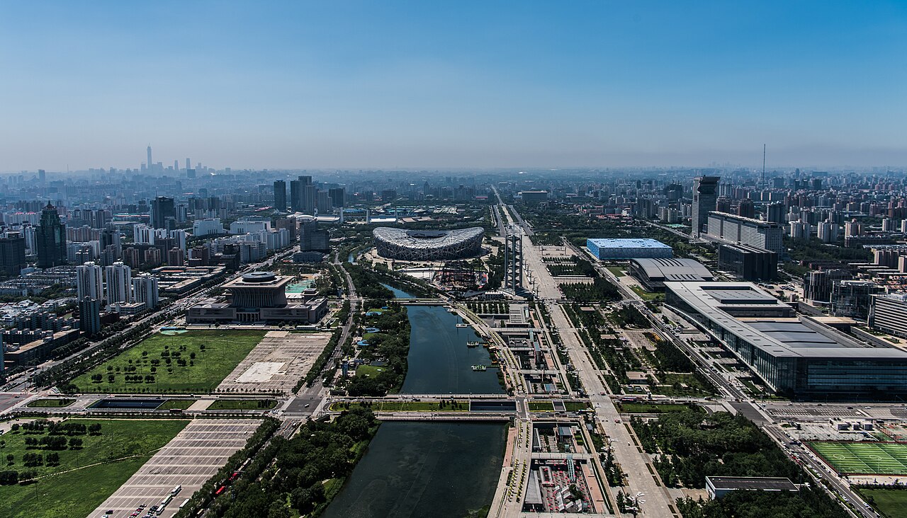 A raised view over the Olympic Green with a long water channel running between lawns and large buildings, and the Beijing skyline beyond