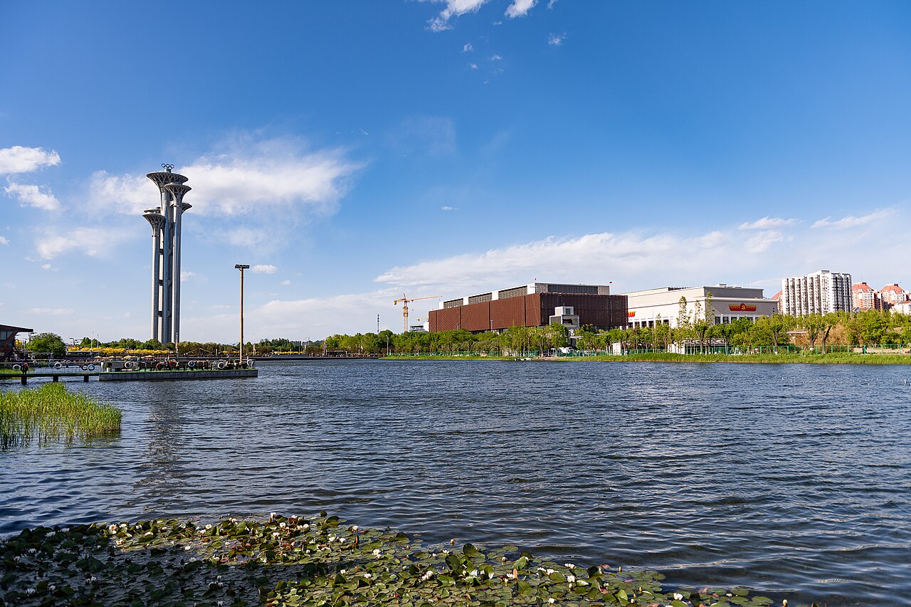 A lake in the Olympic park with reeds at its edge, a tall slender tower with a viewing deck on the far shore and low buildings beside it under a bright sky