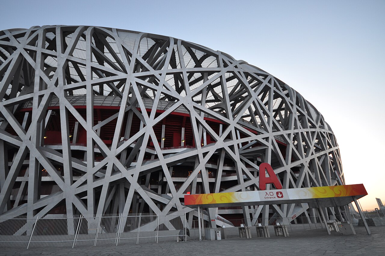 A close view of the stadium's grey steel lattice, with members of different thickness crossing at changing angles and a red and yellow sign low on the structure