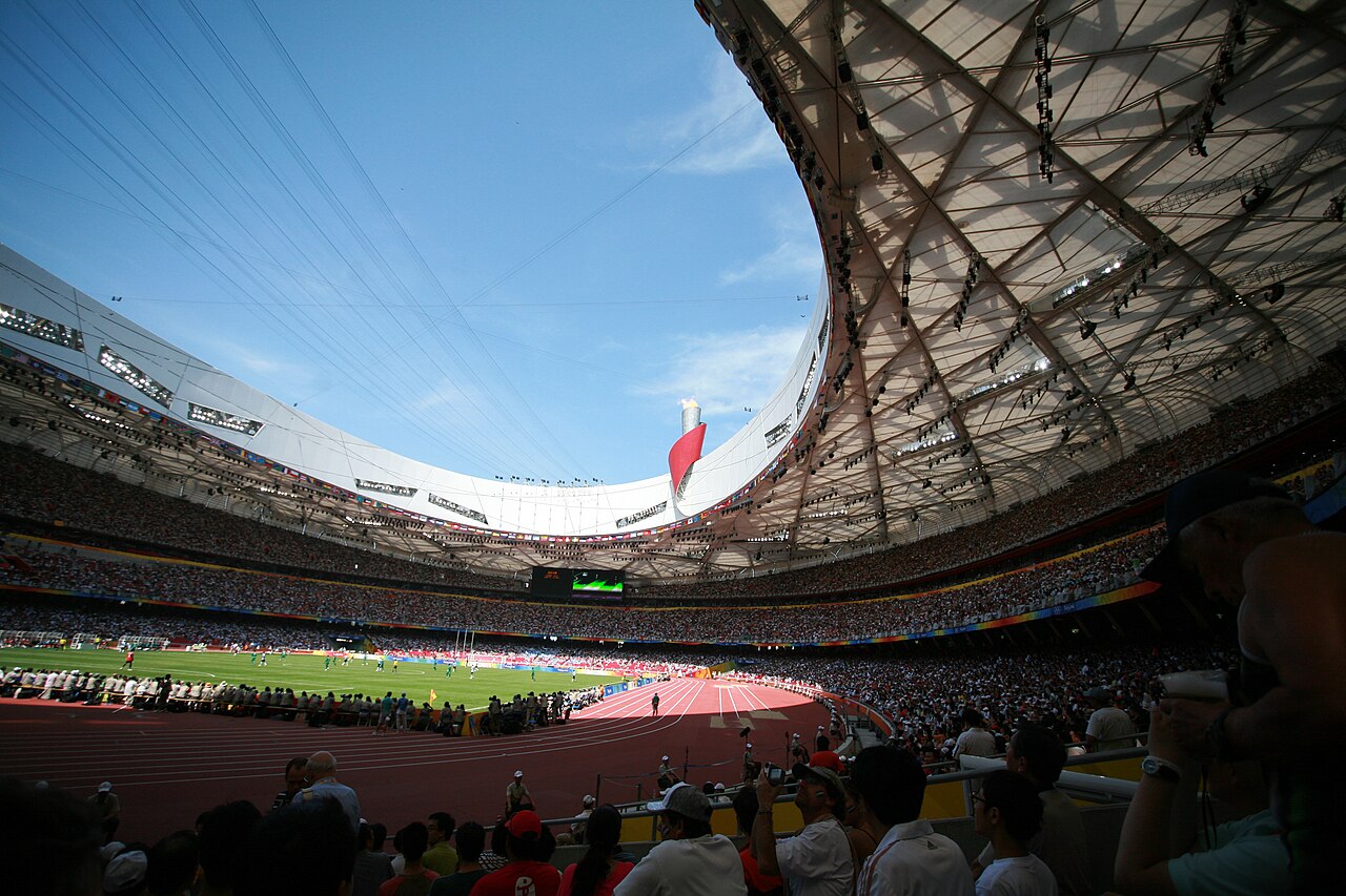 The inside of the stadium roof, a curved ring of steel lattice opening onto the sky above a running track and seating