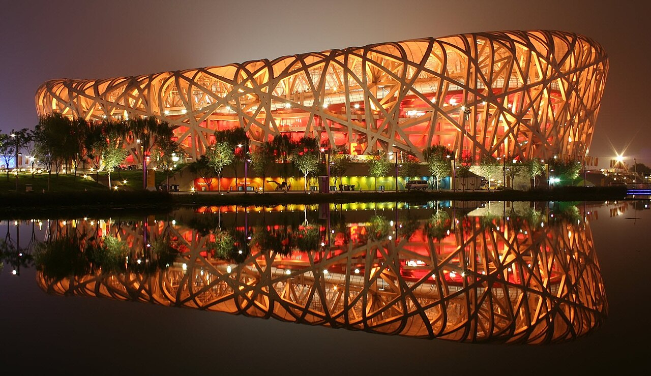 The National Stadium lit orange from inside at dusk, with the whole lattice reflected in still water in the foreground