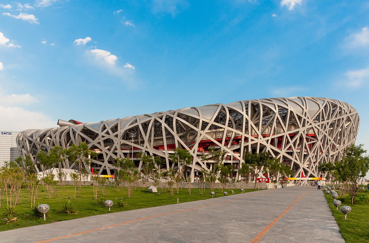 The steel-lattice Beijing National Stadium seen from a paved walkway across lawn and young trees on a bright day, with its grey members crossing the full length of the curved facade under a blue sky with scattered cloud